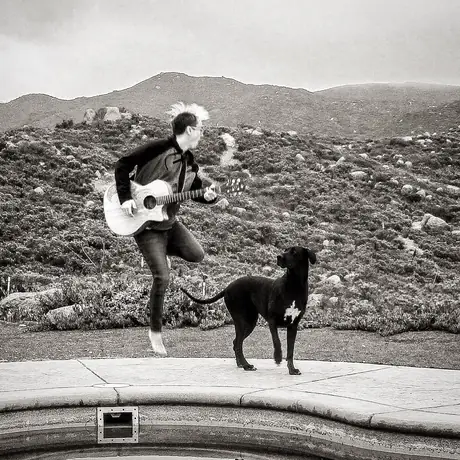 Black and white: a guitarist leaps mid-air on a patio with an acoustic guitar, hair flying, while a black dog stands watching, scrub hills behind.