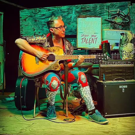 A guitarist plays seated on a small stage lit hard green and pink, an amp beside them and a hand-lettered sign reading Share your talent on the wall.