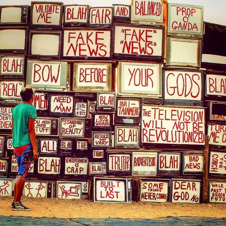 A person walks past a two-storey wall built from old television sets, each screen hand-painted with slogans: Fake news, Bow before your gods, The television will not be revolutionized.