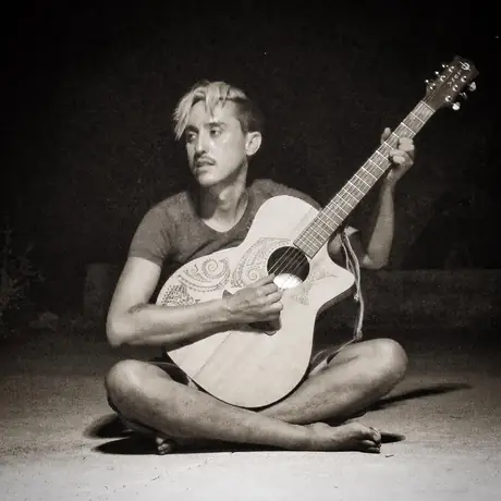 A black and white studio portrait: a musician sits cross-legged on the floor cradling an acoustic guitar, plain backdrop, looking off camera.