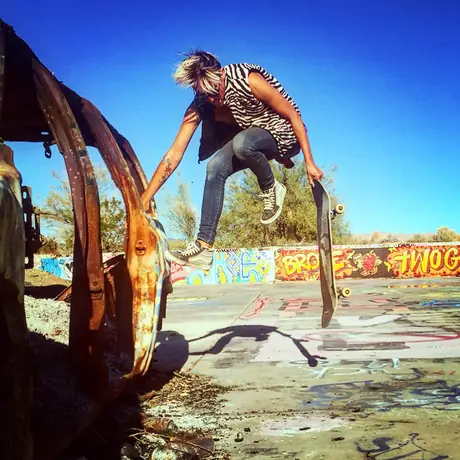 A skater launches off the shell of a burnt-out car in the desert, board underfoot, graffiti across the wreck and hard blue sky above.