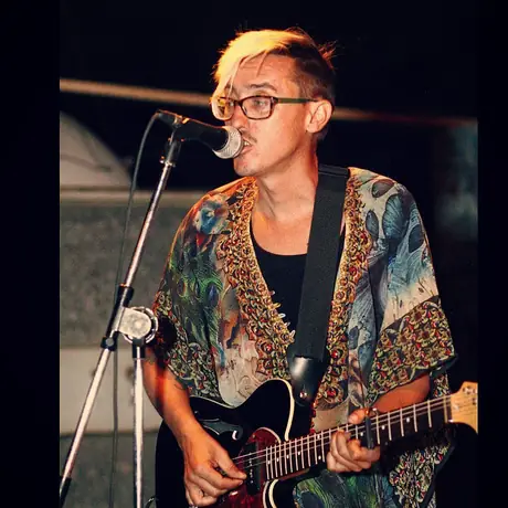 A singer leans into the microphone mid-phrase, electric guitar in hand, wearing a patterned kimono under stage light.