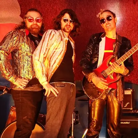 Three musicians pose against a red wall in sunglasses and seventies clothes, one holding a red electric guitar, an amp at their feet.
