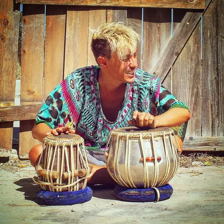A musician sits cross-legged on the ground playing a pair of tabla drums, weathered wooden fencing behind, wearing a bright patterned robe.