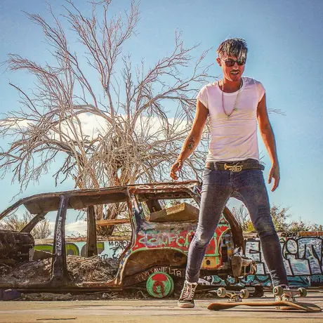 A figure stands on the rusted roof of an abandoned car in the desert, arms loose, a bare tree and graffiti behind.