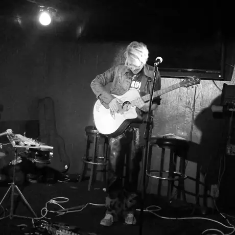 Black and white: a performer plays acoustic guitar seated on a stool under a single lamp in a small dark venue, empty stools in front.