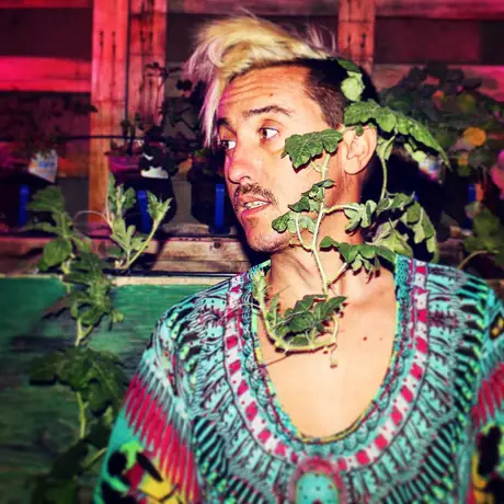 A close portrait with a leafy vine hanging across the face, the subject in a vivid turquoise and pink embroidered top, painted wall behind.