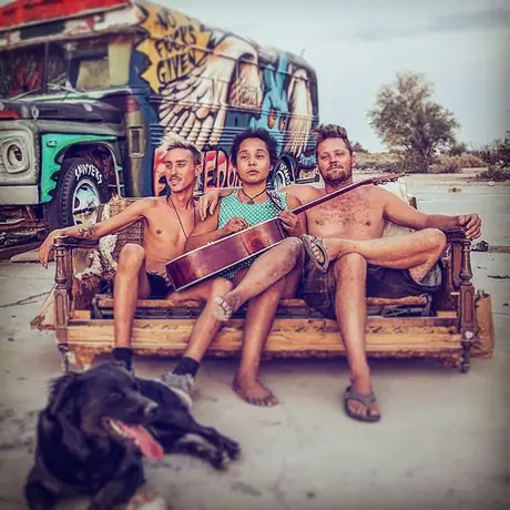 Three people sit on a battered couch outdoors in the desert with an acoustic guitar, a painted bus behind and a black dog lying in front.