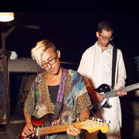 Two guitarists play side by side in a dark room, one in a patterned kimono with a sunburst electric, the other in a white shirt.