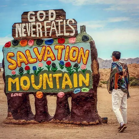 A figure walks past the painted adobe entrance to Salvation Mountain, its hand-made sign reading God never fails above blocks of yellow and red lettering.