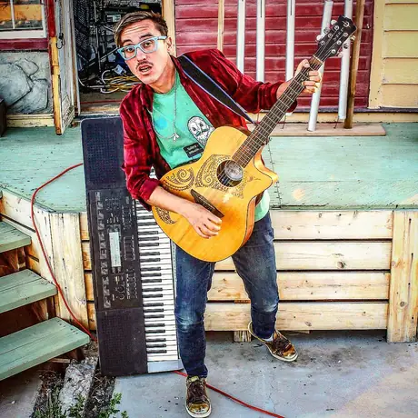 A musician stands playing an acoustic guitar beside an electric keyboard set on concrete steps, red jacket, painted clapboard wall behind.