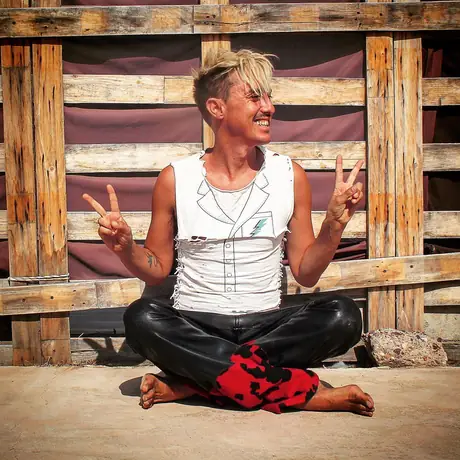 A person sits cross-legged and barefoot on concrete throwing peace signs and laughing, a wall of stacked wooden pallets behind.