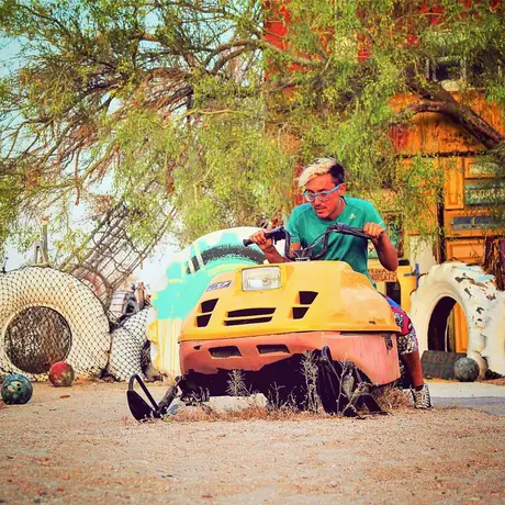 Someone sits on a yellow car body half-buried in desert sand, surrounded by painted tyres and scrap, a shack and tamarisk trees behind.