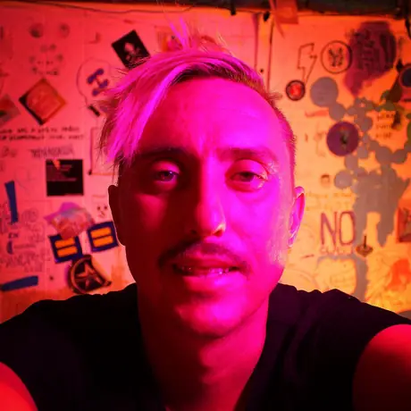 A close self-portrait lit hot magenta, a wall densely covered in stickers and flyers filling the background.