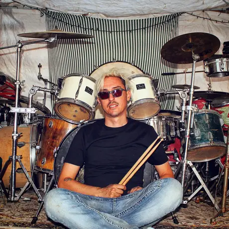 A drummer sits cross-legged on the ground in front of their kit holding a pair of sticks, striped awning behind, sunglasses on.