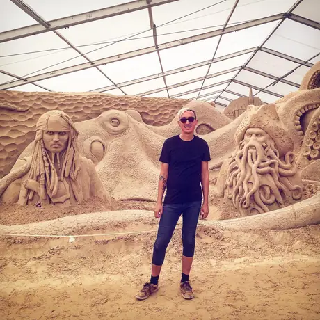 A person stands in front of enormous carved sand sculptures of faces and tentacles under a white marquee.