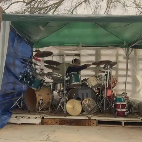 An empty drum kit stands ready under an open-sided canopy on a wooden stage, cymbals and a second kit crowded behind.