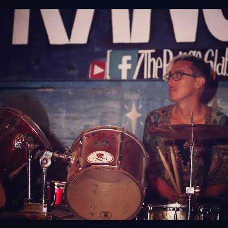 A drummer plays behind a bass drum painted with a skull, a large mural and social handles on the wall behind.
