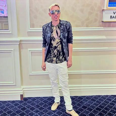 A figure in mirrored visor sunglasses, a marbled blazer and white jeans stands against the panelled wall of a hotel conference corridor.