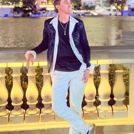A figure in white jeans and an open patterned jacket leans on an ornate gold balustrade in a warmly lit hotel interior.