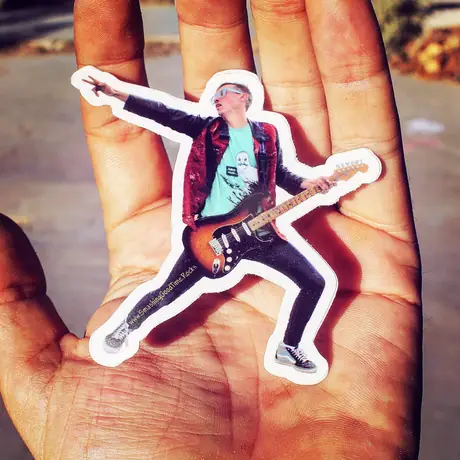 An open palm holds a die-cut sticker of a guitarist mid-lunge, the fingers of the hand filling the frame behind it.